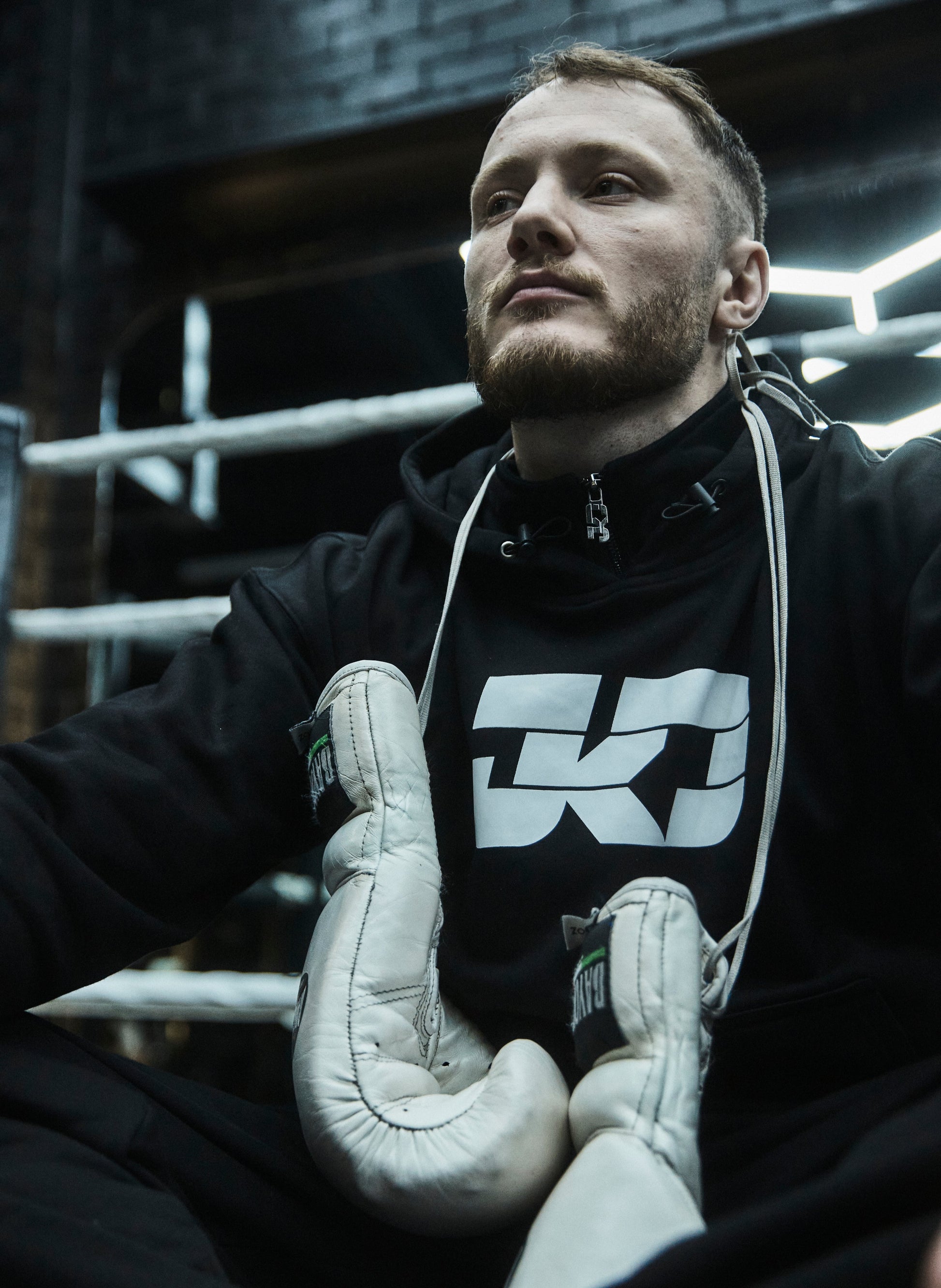 Man wearing a black hoodie with a logo and white boxing gloves in a boxing ring.