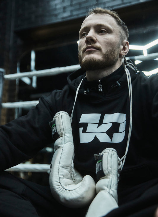 Man wearing a black hoodie with a logo and white boxing gloves in a boxing ring.